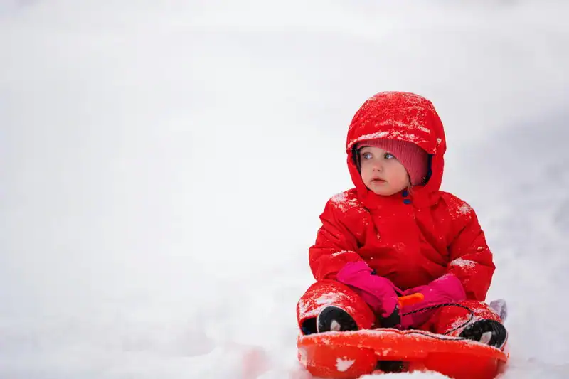 Adorable toddler in red winter gear sitting on a sled in the snow.